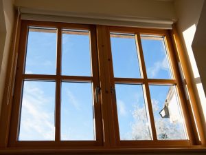 An interior shot looking out through a set of classic wooden-framed windows, with a clear blue sky and some tree branches visible outside, bathed in sunlight.