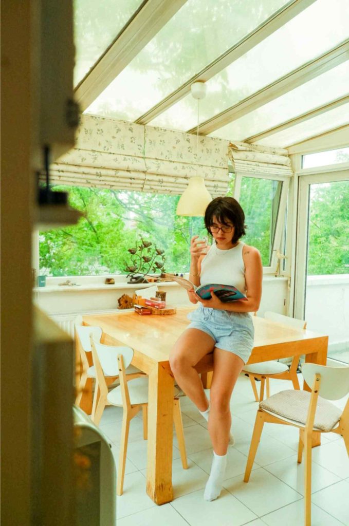 Young woman reading in a sunlit glass sunroom with wooden dining table