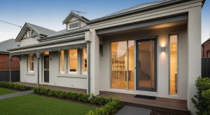 Exterior of a modern and traditional house, featuring a light beige and dark grey color palette, with contemporary floor-to-ceiling glass doors and traditional Victorian wrought iron detailing on the front patio. A neat lawn and green plants are in front.
