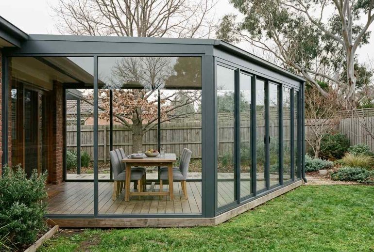 A modern glass-enclosed patio with a wooden deck, featuring a dining table and chairs, surrounded by a lush green lawn and trees.