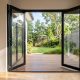 Black aluminium double swing doors open outwards from a living room onto a timber deck and garden, giving a full-width clear opening with no centre track