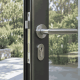 Close-up of a stainless lever handle and euro cylinder on a slim matte charcoal aluminium door, garden visible through the glass